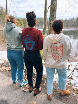 three women standing with their backs towards camera, one in white hoodie with orange design, one in maroon with a blue design and one in a sage hoodie with white design