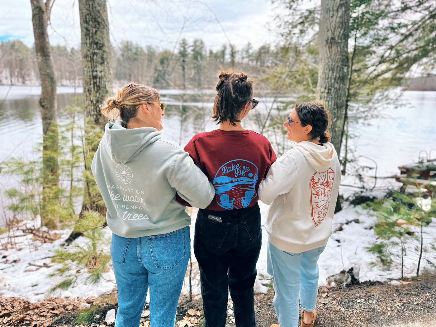 Three women standing in front of a lake looking at each other, wearing three different sweatshirt designs. One in a white hoodie with east coast design, one with maroon in a blue lake life design, and one in a sage hoodie with white water and tree design.